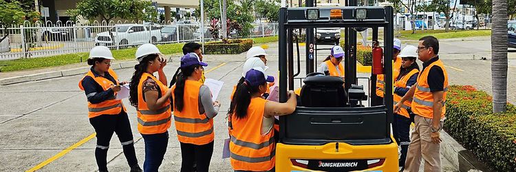 Photo of a group of women wearing safety vests and helmets, standing around an industrial truck while receiving a briefing.