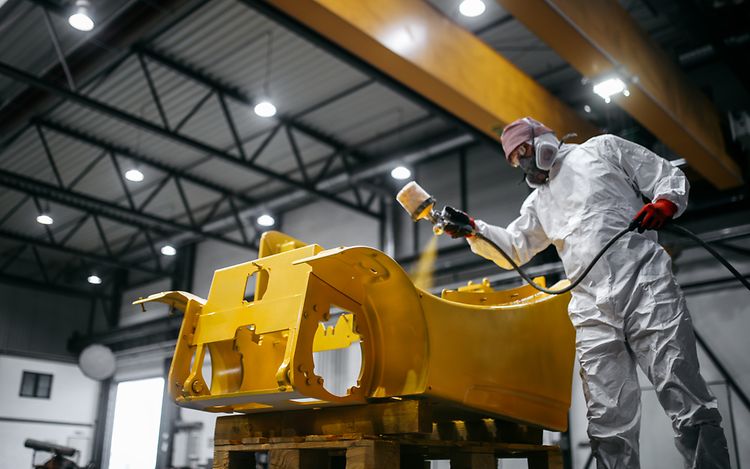 Photo shows a Jungheinrich employee in a protective suit and respirator mask refurbishing a truck with yellow paint. 
