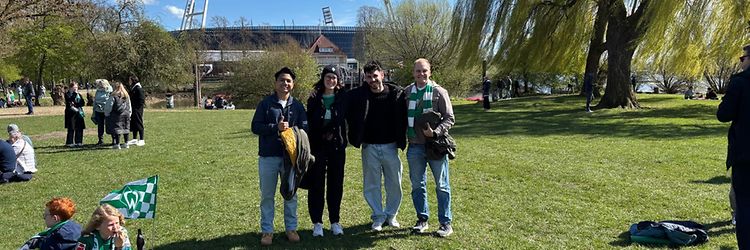 Four people are standing in a field in front of a football stadium, looking at the camera.