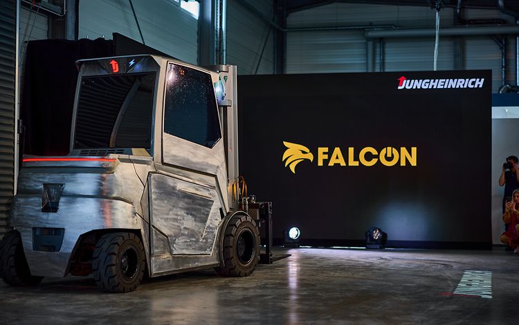 A grey forklift truck is parked in front of a black screen bearing the yellow lettering ‘Falcon’.