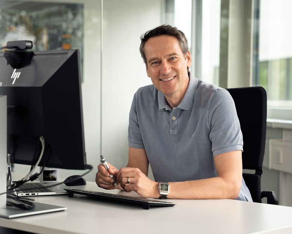 Dr. Tobias Harzer sitzt an einem Schreibtisch und schaut lächelnd in die Kamera.