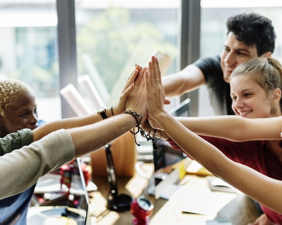 Eine Gruppe von sieben Personen steht in einem Büro und alle legen in der Mitte ihre Hände übereinander und lächeln.