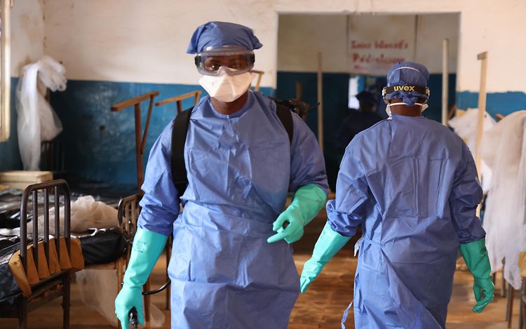 The picture shows two people wearing medical protective clothing. One of them is walking straight towards the camera.