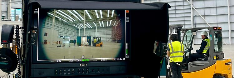 In the foreground is a screen with a script hanging from it. A yellow forklift can be seen in the background.