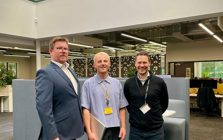 Three men are standing in an office, looking straight at the camera.
