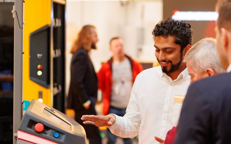 Uzair Mukadam steht neben einem autonomen Mobile Robot und erklärt weiteren Personen etwas dazu.
